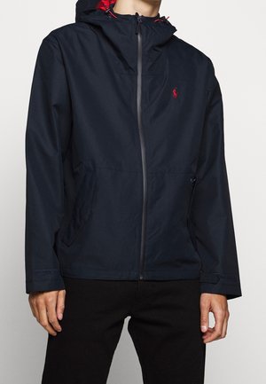 Mann trägt eine marineblaue Jacke mit Reißverschluss, rotem Logo auf der Brust und rotem Kapuzenfutter, kombiniert mit schwarzen Hosen, vor weißem Hintergrund.