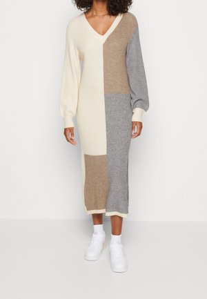 Kvinde iført en lang, farveblokstrikket kjole med creme-, beige- og gråpaneler, kombineret med hvide sneakers og sokker.