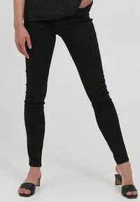 Fekete skinny farmernadrág sima textúrával és szoros illeszkedéssel, fekete, nyitott orrú sarkú mules-szal párosítva, hangsúlyozva a minimalista dizájnt.