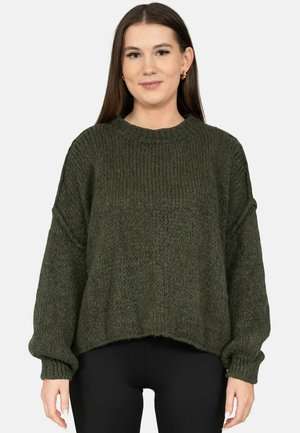 Frau trägt einen locker sitzenden, dunkelgrünen Strickpullover und schwarze Hose, steht vor einem schlichten hellen Hintergrund und blickt nach vorne.