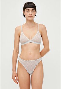 Woman standing against plain background wearing sheer white lingerie set with delicate dot pattern and ruffled edges on panties.
