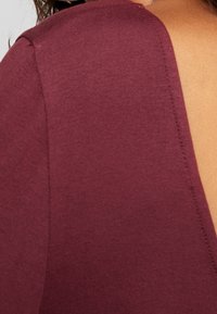 Top bordeaux con scollatura rotonda e design con schiena aperta. Tessuto liscio e texturizzato con dettagli di cucitura lungo i bordi.