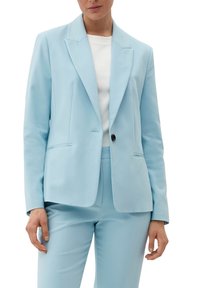 Blazer su misura azzurro chiaro con rever appuntito, chiusura a un bottone, due tasche frontali e texture del tessuto liscia, indossato sopra una camicia bianca.