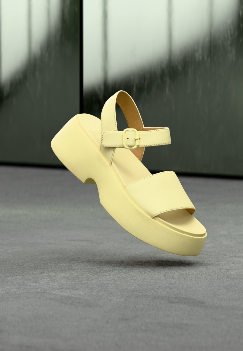 Πλατφόρμα σανδάλι σε ανοιχτό κίτρινο με λουράκι στον αστράγαλο και αγκράφα, που εμφανίζεται στον αέρα με φόντο λείο σκοτεινό τοίχο και γκρι πάτωμα.