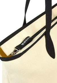 Borsa tote in tela di colore crema, con dettagli neri, chiusura superiore con zip e un'etichetta con logo. Texture liscia e design strutturato.