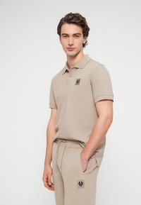 Νεαρός άνδρας με μπεζ πόλο πουκάμισο και ταιριαστό παντελόνι με κονκάρδες λογότυπου, ποζάρει μπροστά σε απλό λευκό φόντο.