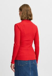 Rotes langärmliges Ripp-Top mit einem geschliffenen hohen Kragen. Hat ein figurbetontes Design und strukturierter Stoff. Getragen mit einem Jeansrock.