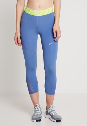 Femme portant des leggings de sport bleu courte avec une ceinture jaune Nike Pro et des chaussures de course grises, debout devant un fond blanc.