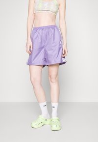 Pantaloncini sportivi viola con fascia elastica in vita e strisce laterali, abbinati a zoccoli verdi e calzini bianchi con dettagli neri.