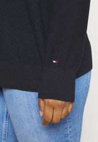Schwarzer Pullover mit strukturiertem Strickdesign. Mit gerippten Bündchen und einem kleinen rot-weiß-blauen Logo auf dem Ärmel. Kombiniert mit blauen Jeans.