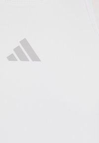 Débardeur de sport blanc en tissu lisse, arborant un logo gris à trois bandes sur le devant. Design élégant avec des détails minimaux.