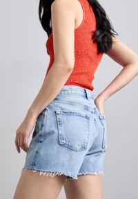 Woman wearing a sleeveless red textured top and light blue denim shorts with frayed hems, posing against a plain light background.