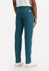 Pantaloni din bumbac teal cu un croi slim, buzunare la spate și un eticheta de marcă roșie pe partea dreaptă. Construiți dintr-un material neted, cu un aspect sofisticat.