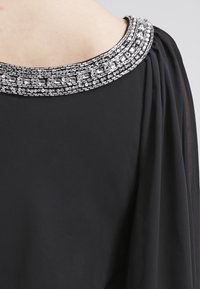 Vêtement noir avec un large col orné, présentant des accents en argent et en cristaux transparents, texture lisse, et tissu fluide qui se drape.