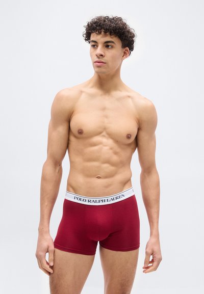 Νεαρός μυώδης άνδρας που φορά κόκκινα μπόξερ της Polo Ralph Lauren, στέκεται μπροστά σε απλό φωτεινό φόντο, κοιτάζει ελαφρώς προς τα δεξιά.