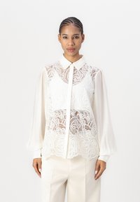 Weiße Bluse mit transparentem Spitzenoberteil und komplizierten Paisleymustern, mit Kragen und langen, gepufften Ärmeln. Glattes Material.