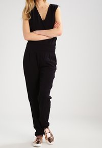 Svart ärmlös jumpsuit med V-ringning, elastisk midja och veck; i kombination med skinande roséguld sneakers. Slät tygtextur genom hela.