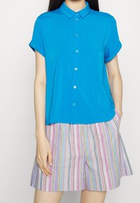 Camicetta blu a maniche corte con colletto, abbinata a pantaloni corti multicolori a righe con linee verticali in tonalità pastello.