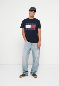 Marineblå bomulds-t-shirt med et rektangulært "TOMMY JEANS" logo-design i rød og hvid. Bæres med lyseblå denimjeans og grønne sneakers.