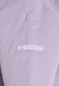 Sudadera de color púrpura claro con textura acolchada, formas redondeadas y una etiqueta blanca con el logo "Freddy" cosida en la parte inferior.