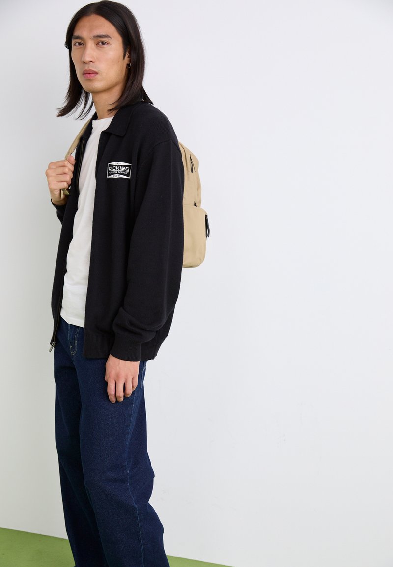 Cardigan noir zippé avec logo brodé, t-shirt blanc en dessous, jeans bleus et un sac à dos beige sur l'épaule droite.