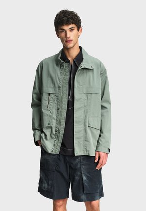 Jeune homme portant une veste boutonnée vert clair par-dessus une chemise noire zippée et un short cargo foncé avec de grandes poches.