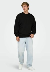 Sudadera negra de algodón con cuello redondo, ajuste holgado, puños y bajos acanalados. Se lleva con vaqueros de corte relajado en azul claro y zapatillas blancas.