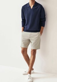Pull marin en tricot bleu marine avec col en V et manches longues, associé à un short beige clair. Des baskets blanches complètent la tenue.