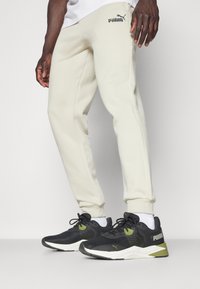 Sorte sportssko med grønne detaljer, parret med lyse beige sweats, der har et lille logo på benet og ribbede ankelmanchetter.