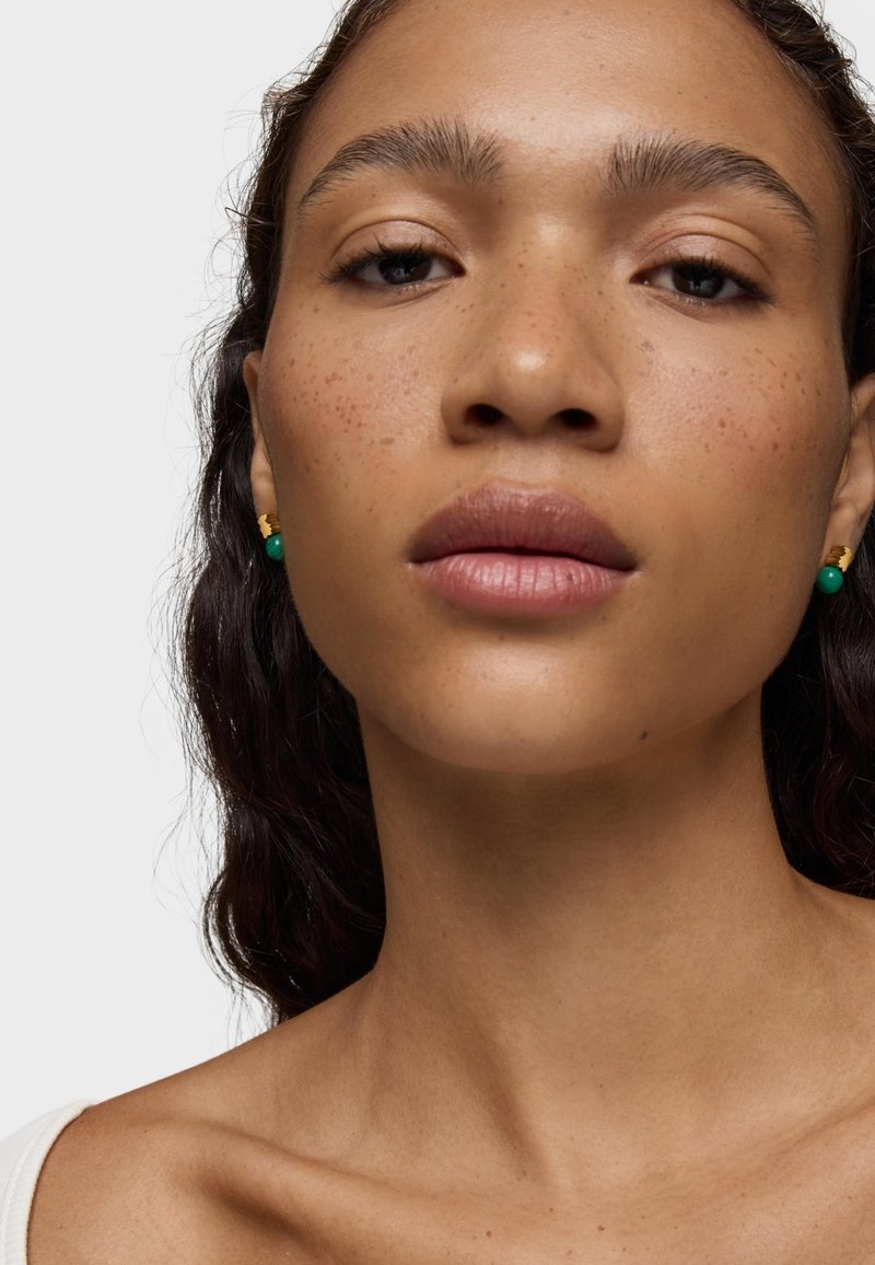 Pendientes dorados y verdes en una persona con piel marrón medio, pecas, labios gruesos y cabello marrón ondulado. Parte de arriba blanca, fondo neutro.