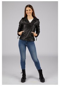 Schwarze Leder-Motorradjacke mit Reißverschluss, seitlichen Taschen und Reverskragen, getragen über einem weißen Hemd und kombiniert mit blauen Skinny Jeans und schwarzen Stiefeln.