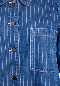 Camisa de mezclilla con rayas blancas verticales, botones de presión negros y un bolsillo en el pecho con detalles de costura en contraste.