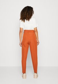 Höga byxor i en bränd orange färg, skräddarsydd passform med sidofickor, bärs med en vit t-shirt. Slät textur och minimalistisk design.