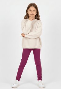 Jasnobeżowy prążkowany sweter dzianinowy i fioletowe legginsy, w białych trampkach. Sweter ma długie rękawy i luźny krój; legginsy są dopasowane.