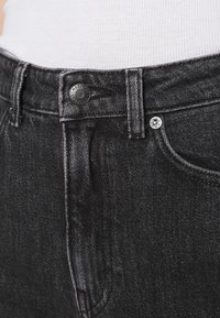 Los jeans de mezclilla negros presentan una cintura alta con un cierre de botón clásico, bolsillos delanteros y una textura sutil debido al tejido de la tela.