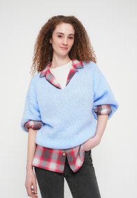 Νεαρή γυναίκα με σγουρά μαλλιά, φορώντας μπλε πλεκτό πουλόβερ πάνω από κόκκινο και γκρι καρό πουκάμισο, ποζάρει με το ένα χέρι στην τσέπη μπροστά σε λευκό φόντο.