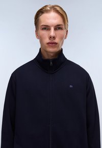 Marineblå zip-jakke med høy krage, laget av glatt stoff. Har en liten logo på brystet og rene sømdetaljer.