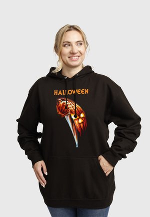 Kobieta ubrana w czarną halloweenową bluzę z kapturem z grafiką krwawego noża i lampionu z dyni, uśmiechająca się i pozująca z rękami w kieszeniach.