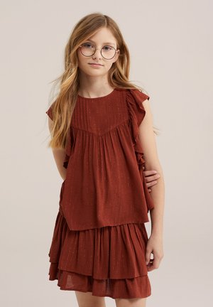 Jeune fille aux longs cheveux blonds et portant des lunettes, habillée d'une robe froncée couleur rouille sur un fond beige uni.