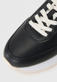 Black leather sneaker with a smooth finish, white laces, and a flat white rubber sole. Features rounded toe and contrasting stitching.