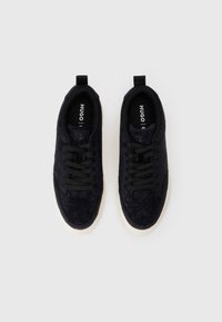 O pereche de adidași negri din suede cu modele brodate subtile, șireturi negre și tălpi din cauciuc alb pe un fundal alb simplu.