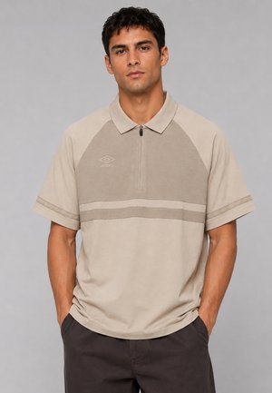 Mann trägt beige kurzärmliges Polohemd mit Reißverschluss und Logo, steht mit Händen in den Taschen vor einfachem grauem Hintergrund.