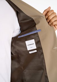 Beige Slim-Fit-Jacke mit einem glatten braunen Innenfutter, versehen mit Etiketten für Marke und Passform sowie einer blauen Akzenttasche. Strukturiertes Design.