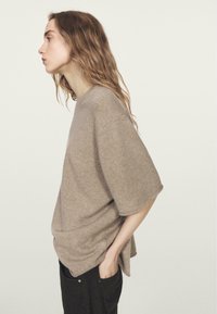 Maglione beige oversize a maglia con maniche corte, collo rotondo e spacchi laterali, abbinato a pantaloni scuri. Tessuto morbido.