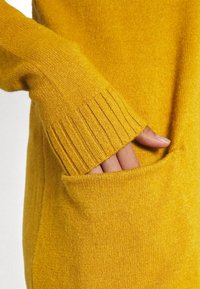 Gul stickad tröja med ribbat ärmavslut och en framficka med en hand som vilar inuti. Mjuk textur med en varm, jämn yta.