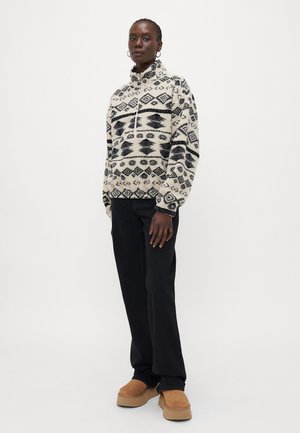 Fleece genser med halvlukking, elfenbenbunn, svarte geometriske mønstre. Kombinert med svarte bukser og beige platåsko.
