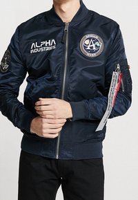 Chaqueta bomber azul marino hecha de nylon brillante. Cuenta con cremallera frontal, parche en el pecho, logo y etiqueta roja en la manga. Puños y cintura acanalados.