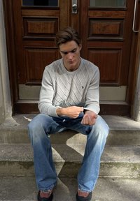 Mladý muž sedí na vonkajších schodoch pred hnedými drevenými dverami, upravuje si rukáv svetlosivého svetra, nosí modré rifle a hnedé topánky.