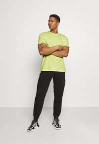 Maglietta verde lime a maniche corte in cotone, abbinata a pantaloni jogger neri. I pantaloni presentano un design sottile. La calzatura è composta da sneakers bianche e nere.
