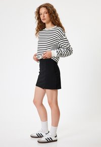 Schwarzer Rock mit einem Schlitz auf der Vorderseite, kombiniert mit einem weißen gestreiften Langarm-Oberteil. Weiße Socken und schwarz-weiße Sneaker runden den Look ab.